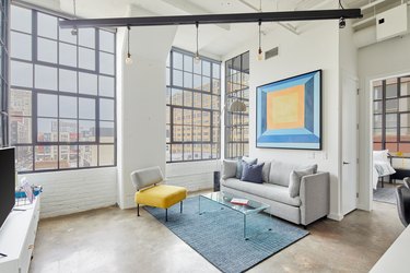 Modern loft living room with concrete floors, gray couch, yellow accent chair, plexi coffee table, brushed metal floor lamp, blue area rug.